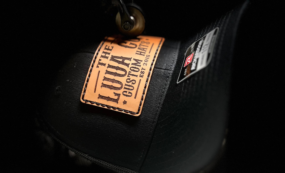 Black cap with a brown leather patch labeled 'The Loua Co. Custom Hats' on a dark background